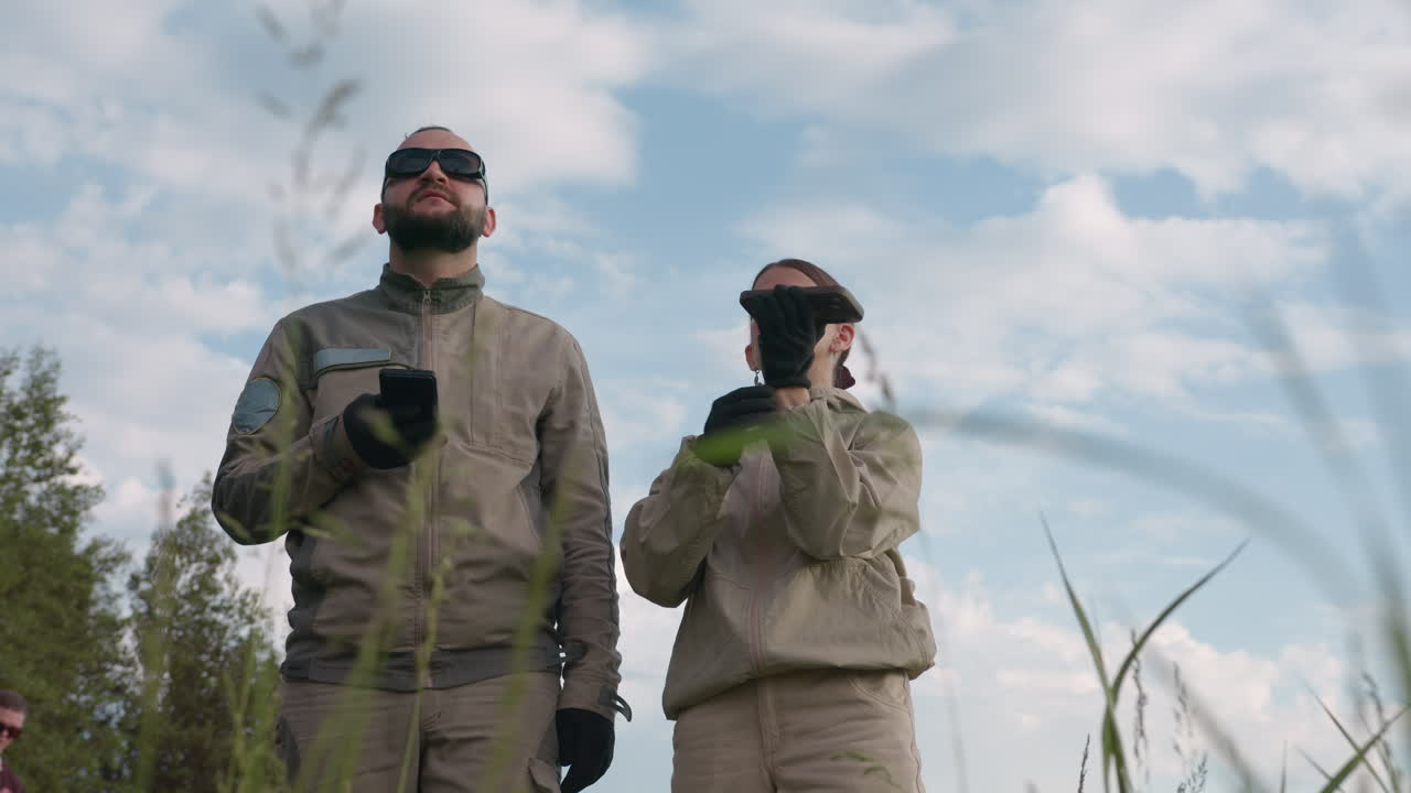 woman pointing remote controller skywards while male colleague in dark sunglasses watches intently through tall grass under cloudy sky, gloved hand adjusts device