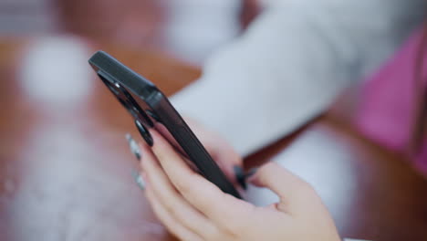 close-up partial view of hand holding smartphone on wooden table with focus on device interaction, black polished nails visible, demonstrating modern mobile use in a stylish setting