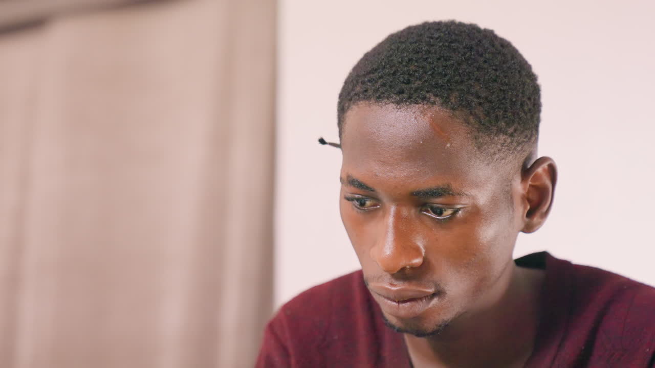 Black man closeup portrait showing concentration, intense gaze toward screen, beads of sweat on forehead, burgundy sweater, neutral curtain backdrop, cinematic soft lighting, internal debate