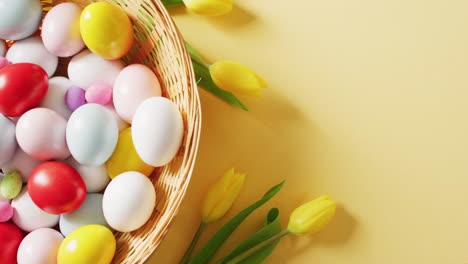 Basket-with-colorful-easter-eggs-and-tulips-on-yellow-background-with-copy-space