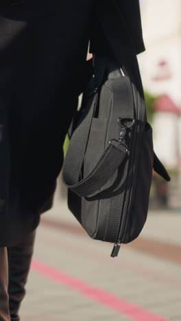 vista trasera de una mujer caminando con confianza en una calle urbana iluminada por el sol, con un elegante abrigo negro y elegantes botas de cuero, llevando dos bolsas, el fondo borroso muestra a otra persona caminando con una chaqueta rosa