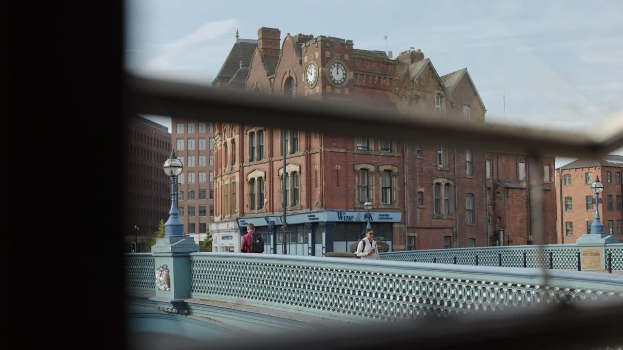 Urban commuters walk across city bridge, viewed through railing, with soft daylight and steady camera