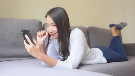 young happy asian woman lying on couch and looking at smartphone, social network or selfie picture concept, full frame slow motion