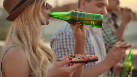 una donna mangia pezzi di pizza calda e beve birra da una bottiglia di vetro verde alla festa con il suo amico in una calda giornata d'estate.