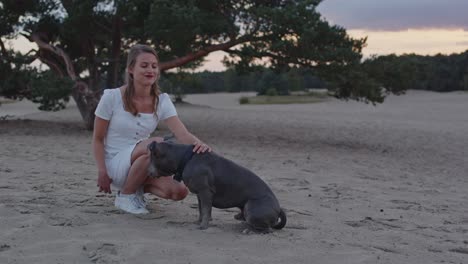 mujer joven acariciando a su perro american staffordshire terrier en dunas de arena y sonriendo a la cámara