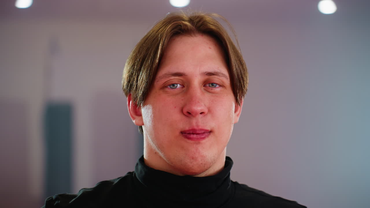 Male in black turtleneck stands in gym facing camera with serious and confident expression, illuminated by strong lights, embodying strength, discipline, focus, and readiness for intense training