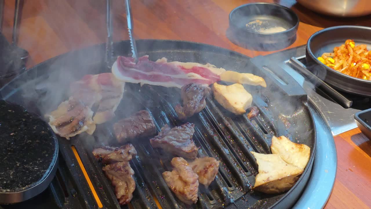 Hands grill marbled beef slices and king oyster mushrooms on a hot tabletop Korean BBQ grill with side dishes