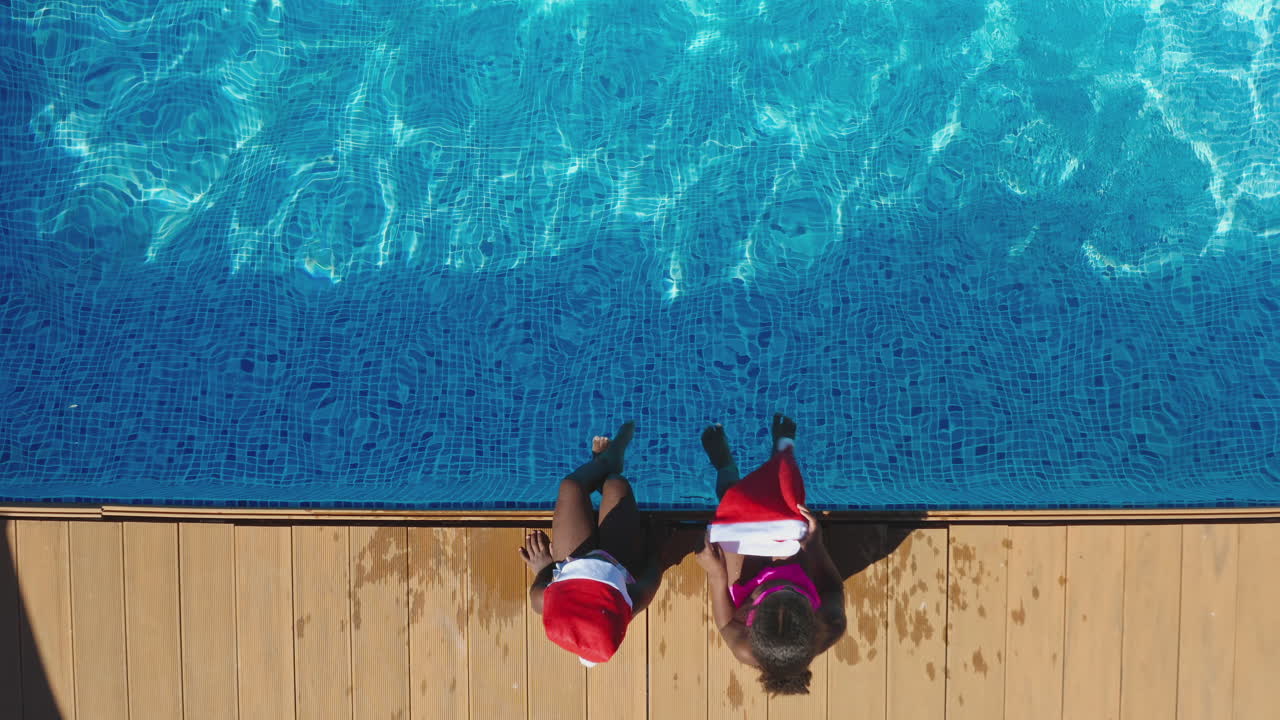 Foto Drone Keluarga Multi-Generasi Saat Liburan Natal Duduk di Tepi Kolam Renang Mengenakan Topi Santa