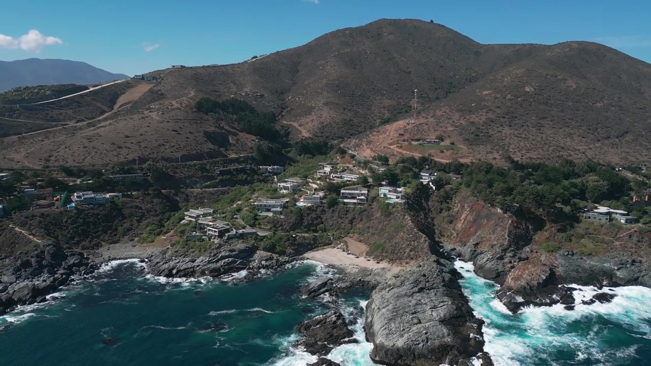 Revelação panorâmica recuando sobre villas exclusivas no topo de falésias com piscinas integradas na encosta de matagal acidentado, acima de uma enseada rochosa isolada e ondas a rebentar. Punta Pite, Papudo, Chile.