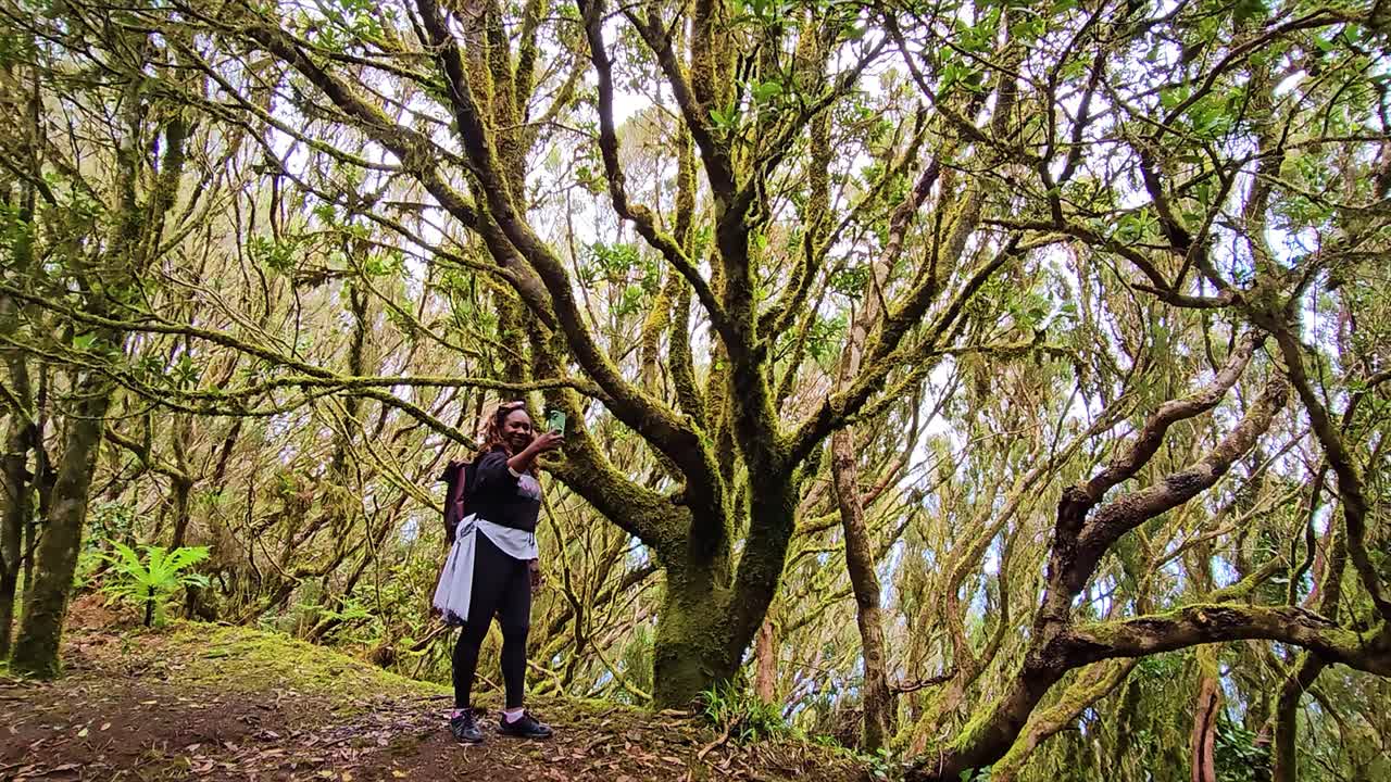 Exploring lush forest trails, a woman captures a scenic shot amidst moss-covered trees in Anaga, Spain