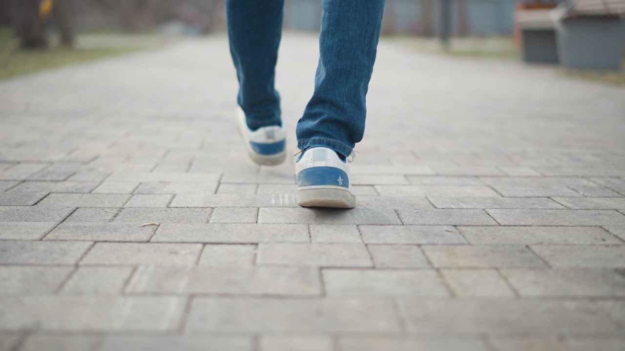 Close-up of person walking on paved path wearing blue jeans and sneakers, capturing motion and footsteps from behind with focus on shoe sole, suggesting journey, solitude, progress, or urban daily routine