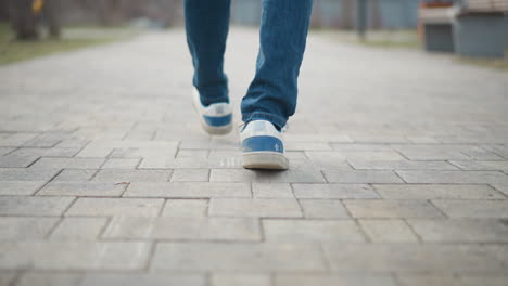 close-up of person walking on paved path wearing blue jeans and sneakers, capturing motion and footsteps from behind with focus on shoe sole, suggesting journey, solitude, progress, or urban daily routine