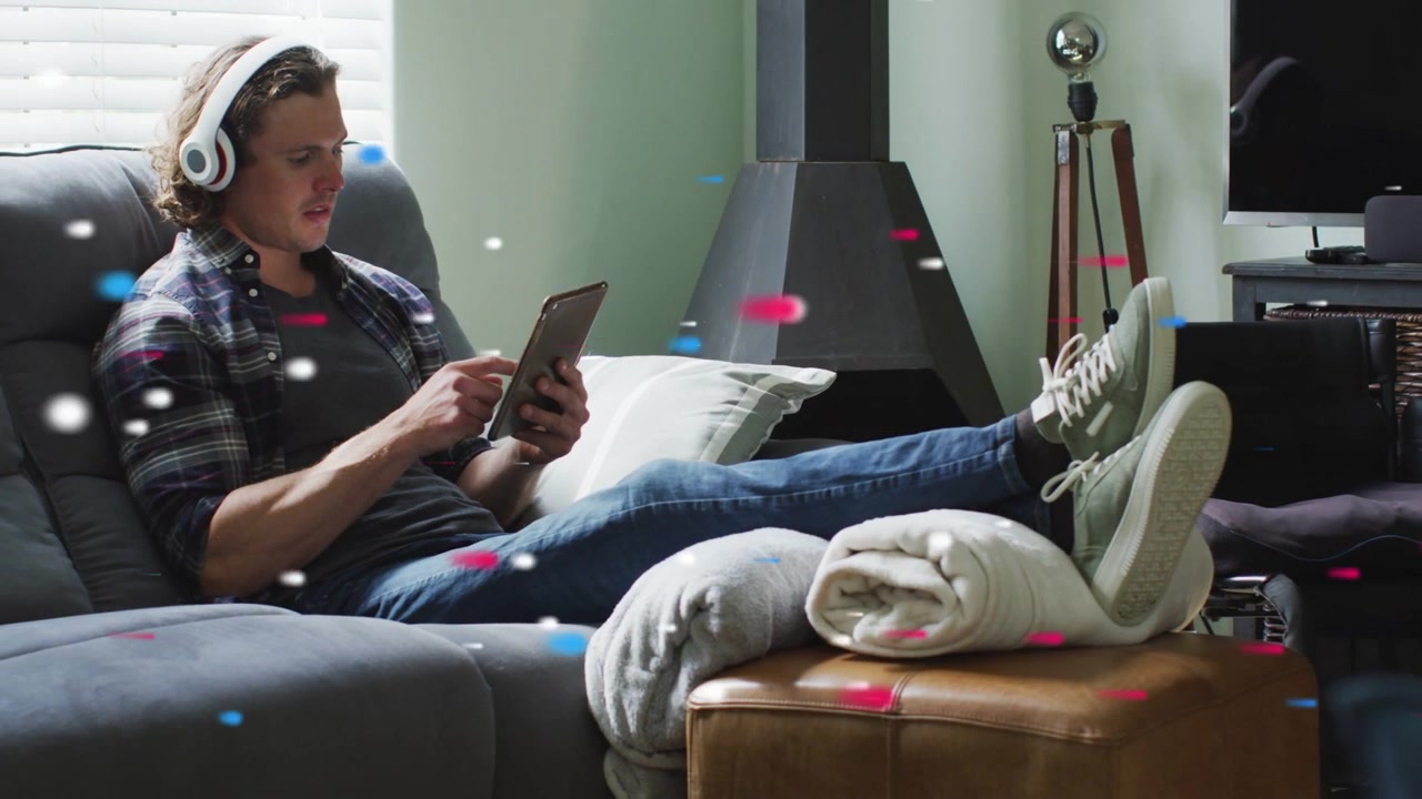 Man reclining on sofa tapping tablet, headphones on, streaming tech content, particles overlaying