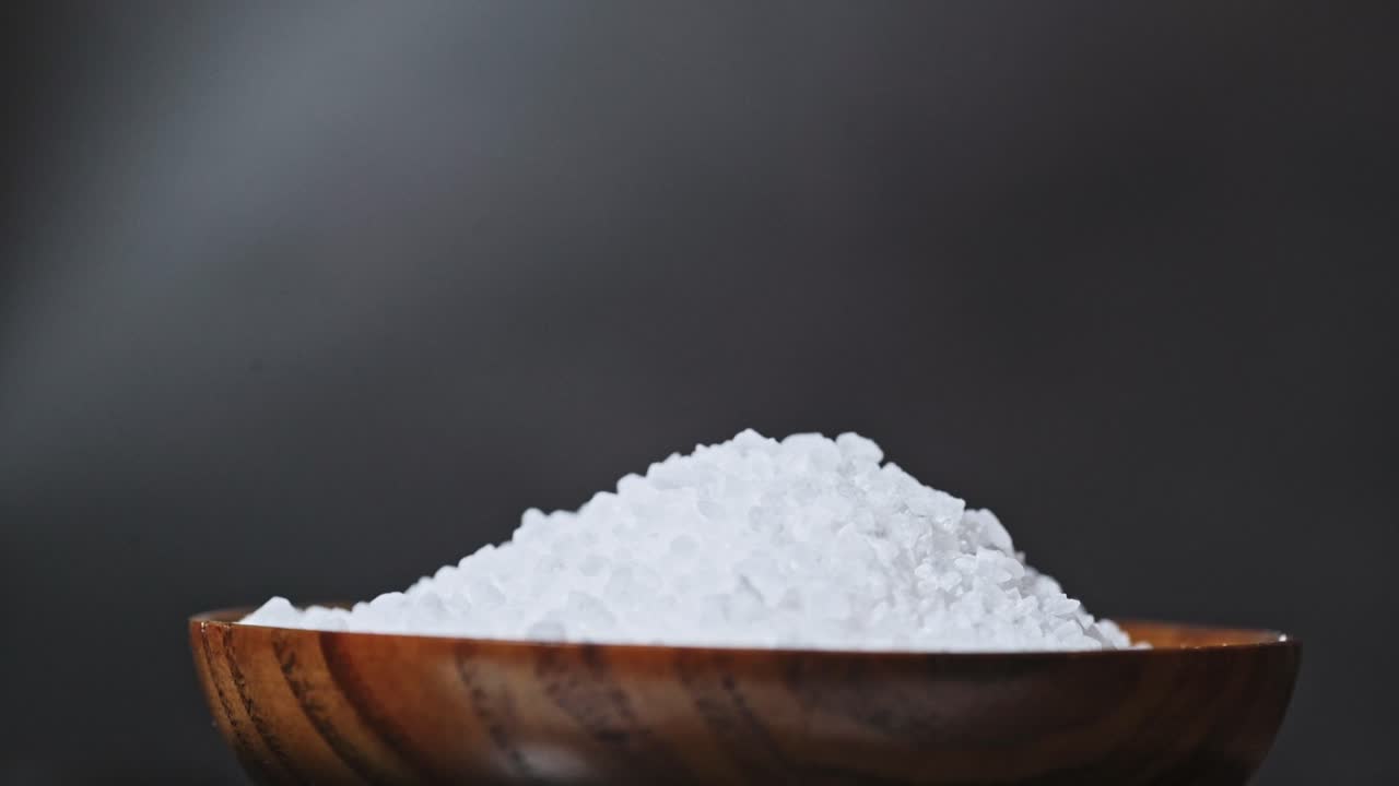 A close-up of a wooden bowl filled with coarse sea salt against a dark, blurred background. The white crystals contrast beautifully with the rich brown tones of the bowl.