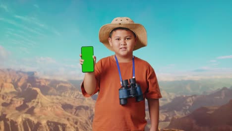 asian boy with a hat and binoculars smiling and showing green screen smartphone to camera. boy researcher examines something while traveling at the top of mountain, travel tourism adventure concept