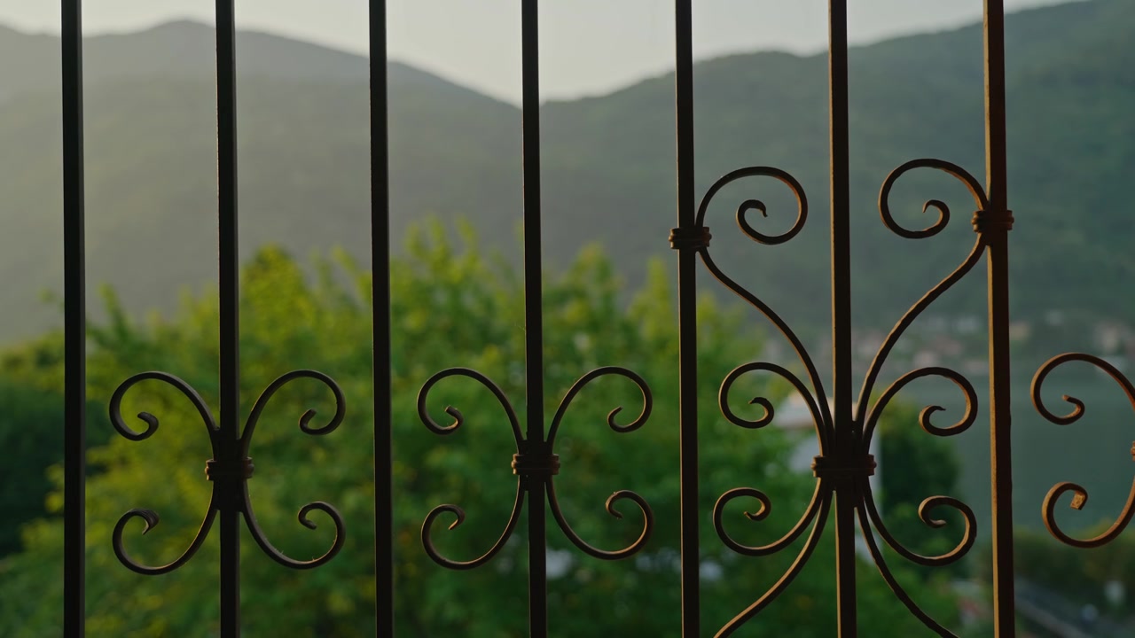 Una prospettiva ravvicinata attraverso un tradizionale balcone italiano ornato, con una balaustra decorativa a fuoco e un bokeh sognante di lago e montagne sullo sfondo, che evoca l'eleganza mediterranea