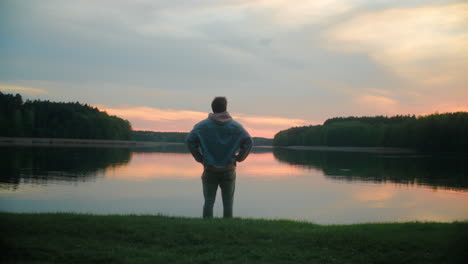A man standing in front of the shore of a Polish lake at sunset. Colorful sky reflects on the water, creating a peaceful, scenic landscape filled with calm and natural beauty