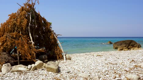 hut with branches and leaves on secret beach with pebbles washed by clear crystal water of ionian sea