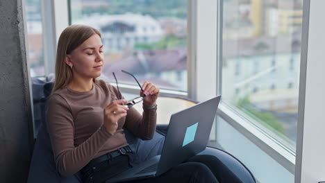 blonde lady takes off glasses finishing work at laptop. tired young woman closes computer and looks into window in front of her smiling.