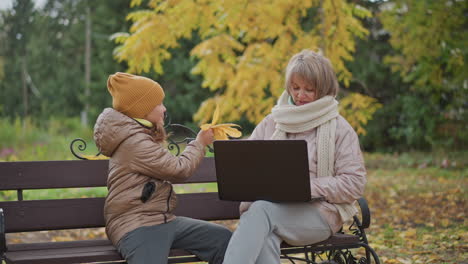 woman sits bench working on laptop outdoors in autumn park while little girl approaches holding golden leaves to distract mother and mother gently shifts hand aside before child moves