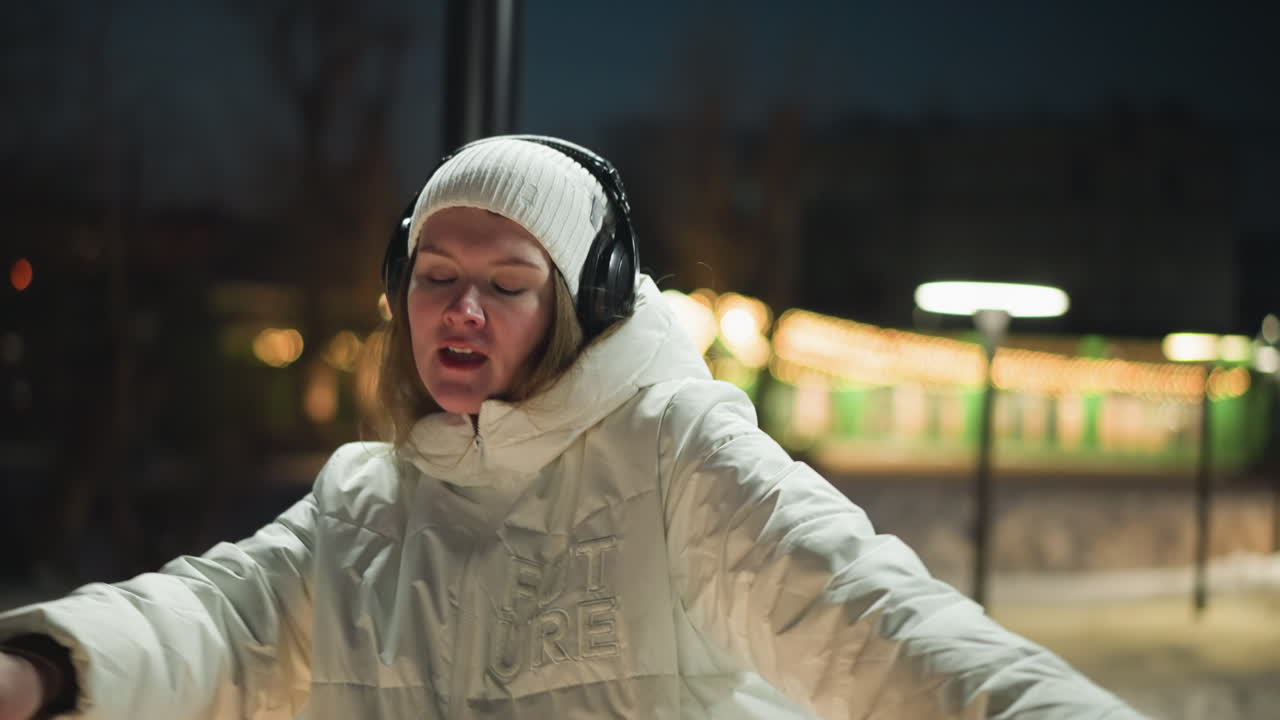 Performer dancing passionately under glowing street lamps on snowy park walkway at night wearing white puffer coat beanie and headphones with eyes closed and joyful expression