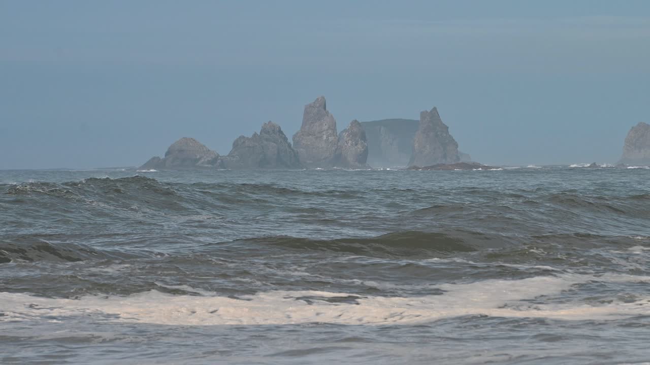 A drone captures waves rolling onto the sandy shoreline of Olympic National Park, Washington. Rocky sea stacks rise from the ocean in the distance under a clear blue sky