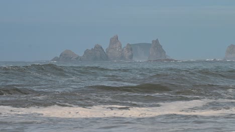 a drone captures waves rolling onto the sandy shoreline of olympic national park, washington. rocky sea stacks rise from the ocean in the distance under a clear blue sky