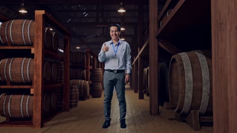 full body shot captures of an asian male professional worker standing in cellar of winery, his broad smile at the camera and giving thumbs up