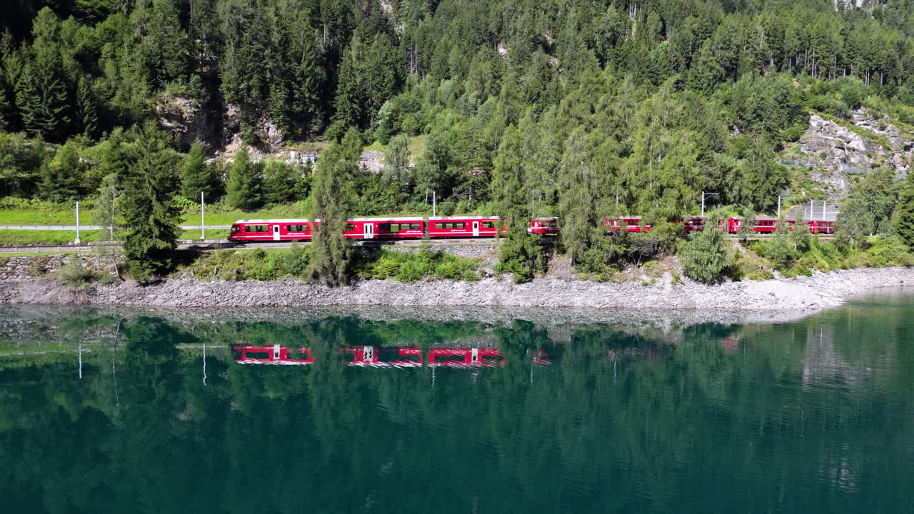 Red train travels by lake with trees reflecting in calm water