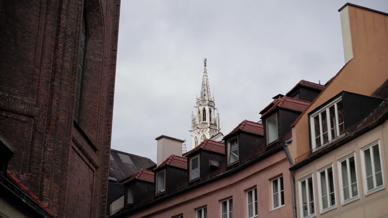 A Gothic-style tower rises above the rooftops of Munich, Germany, framed by historic buildings. The cloudy sky enhances the contrast between the ornate white spire and the warm-toned architecture.