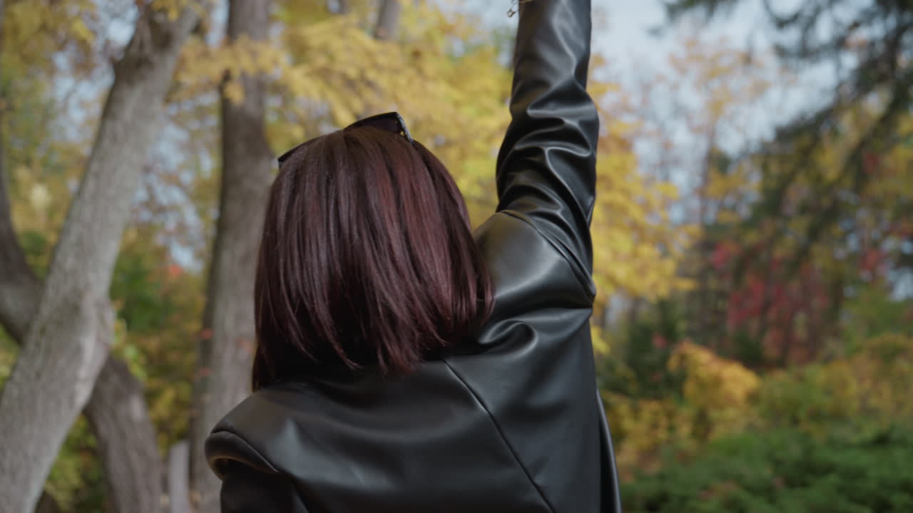 woman in leather jacket facing trees, reaching toward canopy with confident gesture back portrait shows textured hair and studded black jacket against yellow autumn leaves, park grove atmosphere