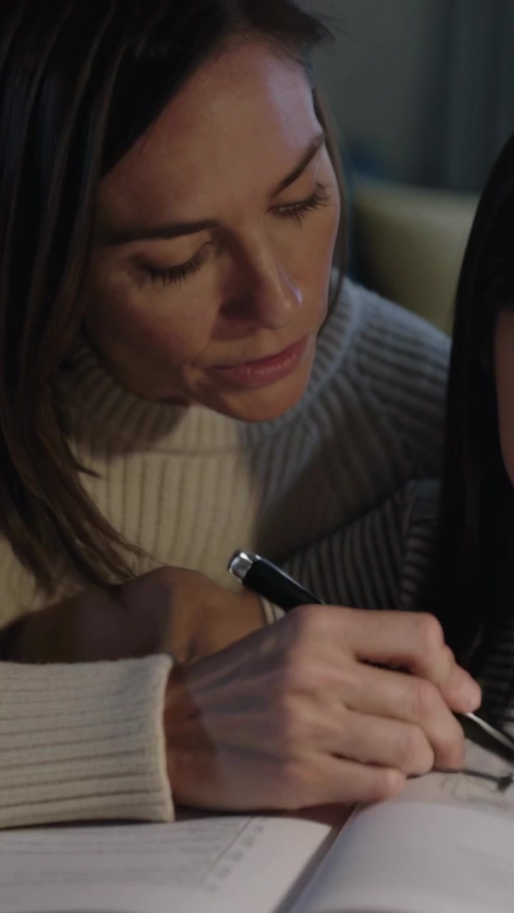 Video verticale: Aprendo un quaderno di esercizi, una donna con un maglione guida un bambino sul divano, indicando con una penna.