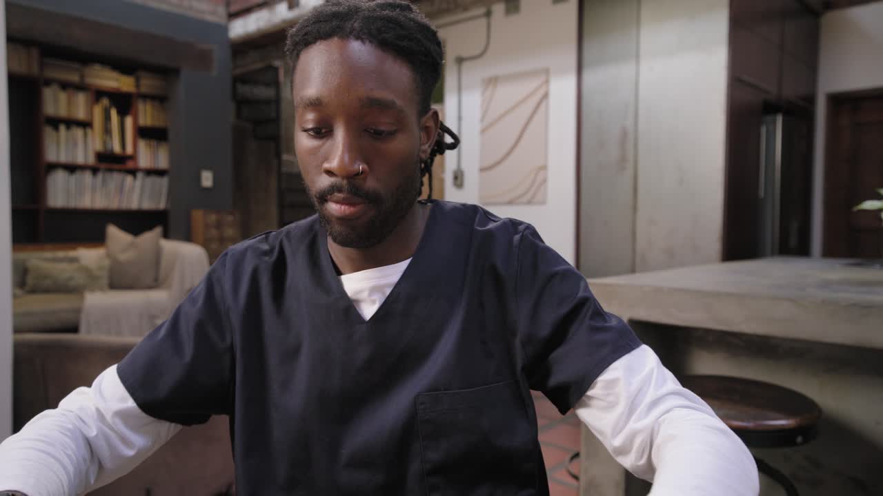 Un uomo africano in camice entra in un loft, tenendo una bottiglia, si siede, regola la telecamera e parla in camera.