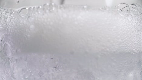 verter soda en cubitos de hielo en macro de vidrio llenándose con burbujas de carbonatación en cámara lenta agua con gas vertiendo de cerca