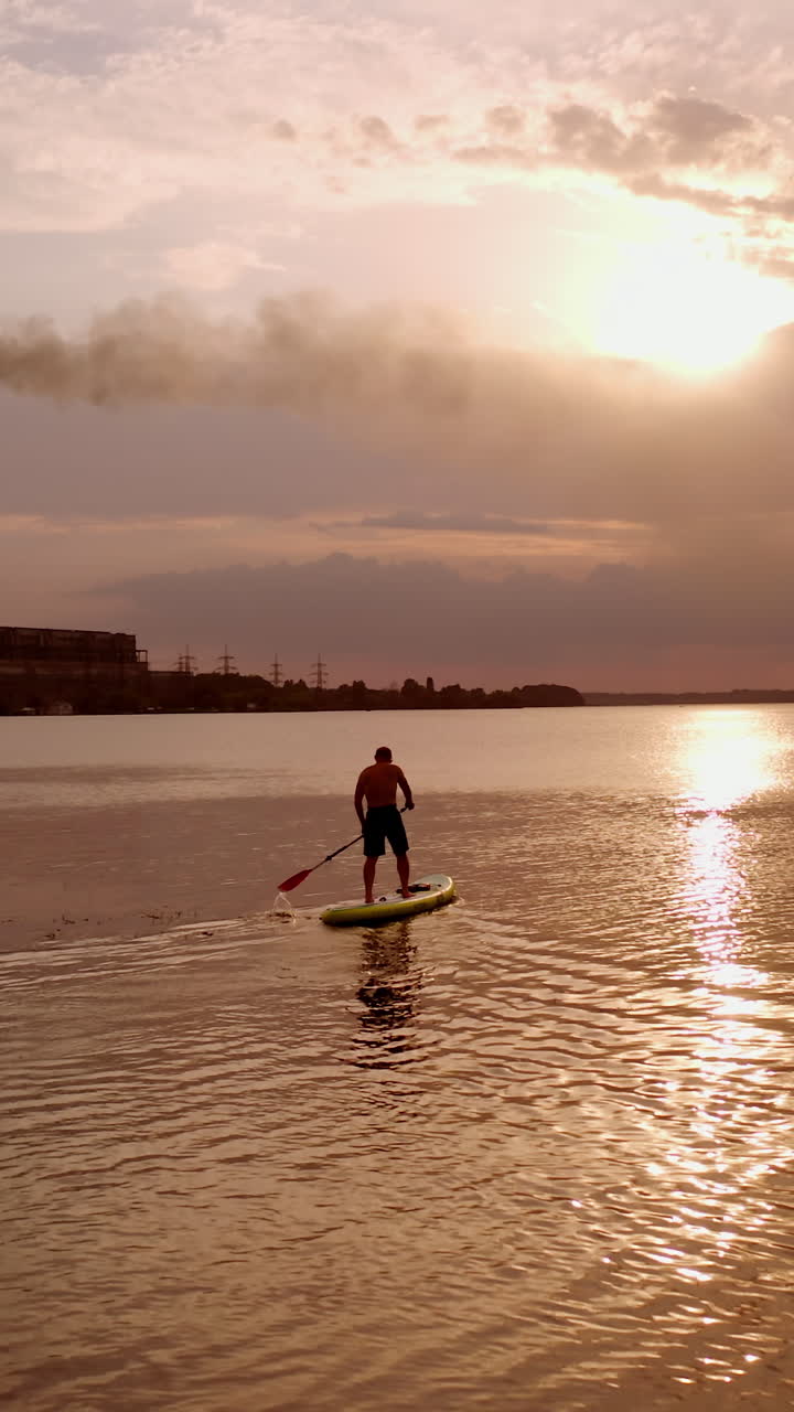 Paddleboarding at sunset. Sportive guy sailing on a board with oars on the evening water on the background of industrial chimneys with smoke. Travel on water. Vertical video
