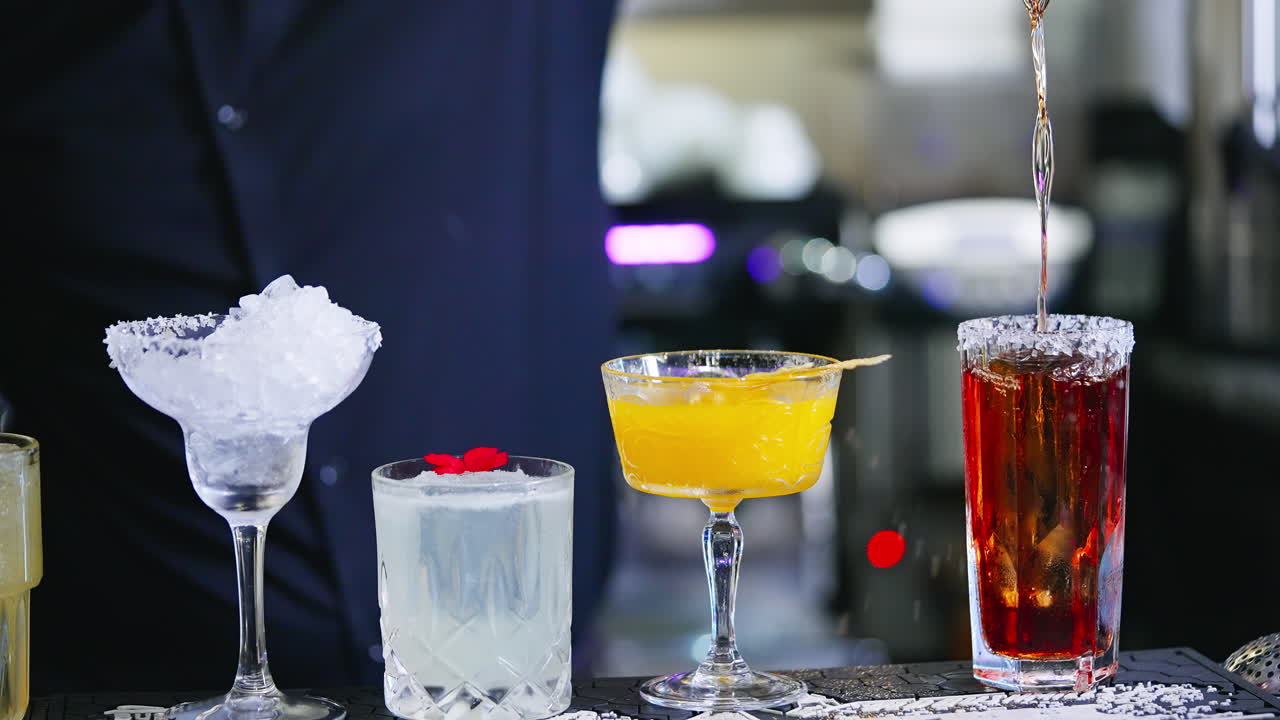 Beautiful tasty cocktails in diverse glasses. Barman in black shirt pours red beverage into a glass with salt on top. Close up.