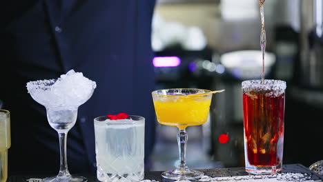 beautiful tasty cocktails in diverse glasses. barman in black shirt pours red beverage into a glass with salt on top. close up.