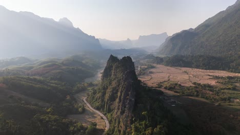 Steep limestone ridges rise from the lush Nam Xay landscape near Vang Vieng, Laos, with a winding road below, patchwork fields, and surrounding mountain ranges bathed in warm atmospheric light
