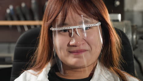 estudiante con escudo de protección facial parpadea los ojos, se ríe con una cálida sonrisa en el taller mecánico, el fondo presenta herramientas industriales, iluminación brillante y maquinaria