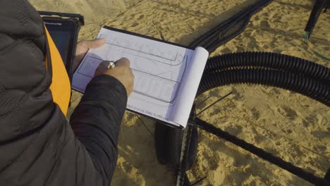 close-up of a man taking notes on paper plans at a sandy construction site with pipes and dark structure - israel