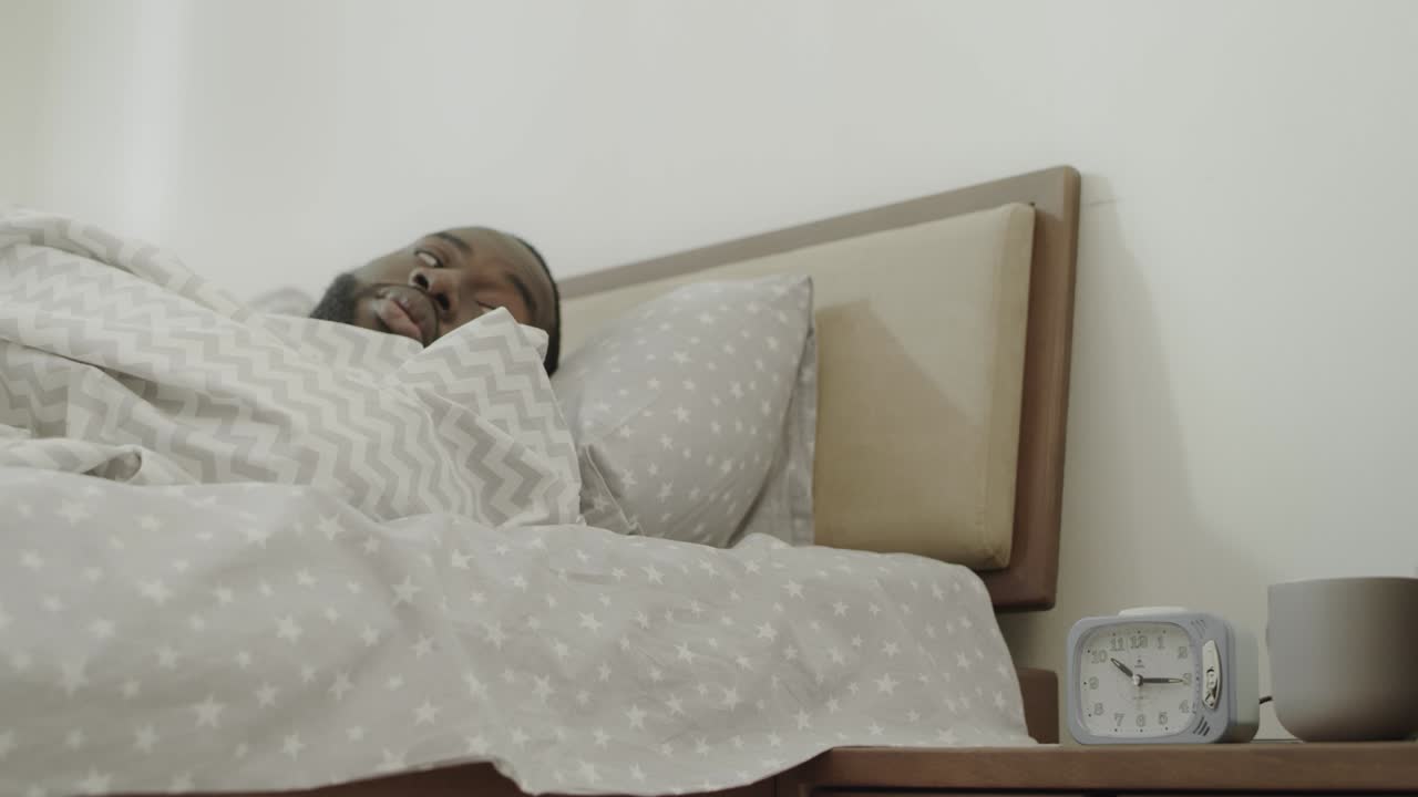 hombre negro apagando la alarma por la mañana. joven adulto despertando en la cama lentamente.