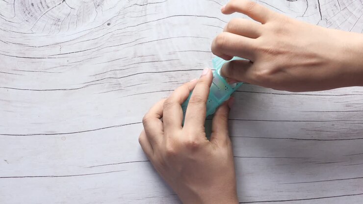 Person opening a light teal pill organizer on a wooden table.