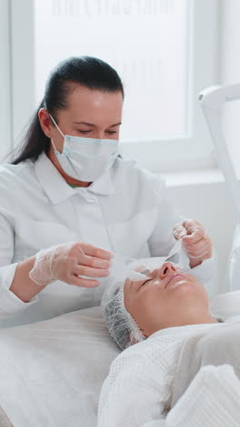 beautician removing regeneration moisturizing sheet mask from young adult woman face skin and eyes