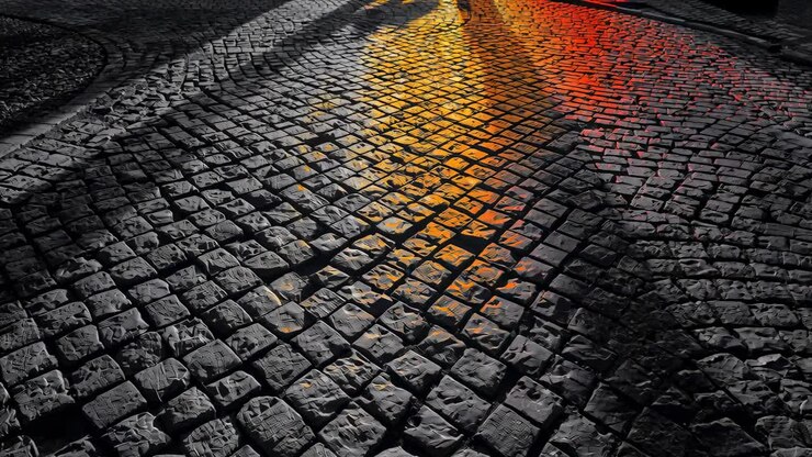 Shifting light bathing cobblestone street with wet patches, expanding central yellow-red reflection