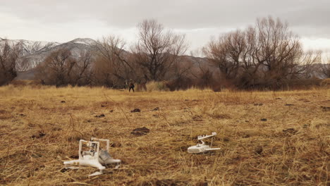 Static-shot-of-white-drone-upside-down-on-the-ground-with-the-controller-in-the-background-a-man-walks-away-then-runs-in-brown-grass-with-snowy-mountains-in-a-cloudy-sky