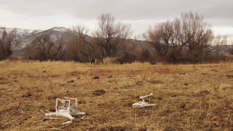 static shot of white drone upside down on the ground with the controller in the background a man walks away then runs in brown grass with snowy mountains in a cloudy sky