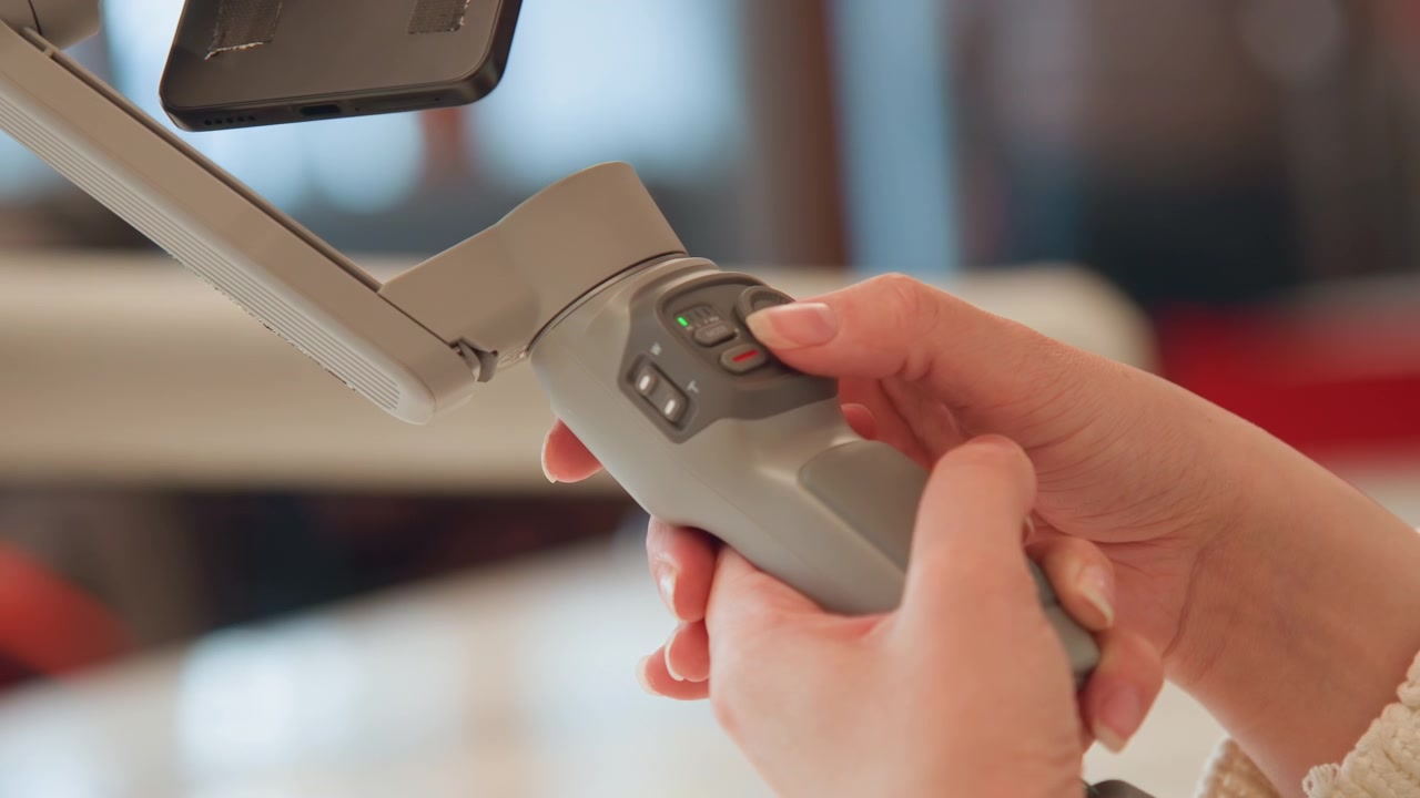 Close-up shows hands actively adjusting handheld gimbal controls, thumb pressing directional pad while fingers maintain steady grip and fine tune stabilization settings on compact device for smooth