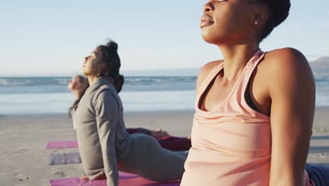 Group-of-diverse-female-friends-practicing-yoga,-stretching-at-the-beach