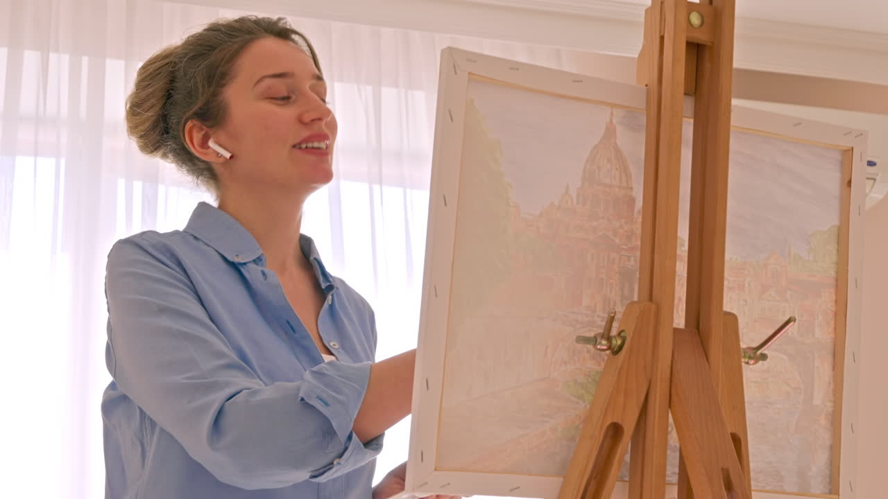 Caucasian woman painting a picture with a palette in her hands and listening music with earphones indoors