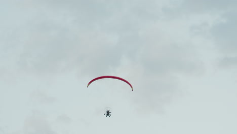back view of paraglider pilot soaring into pale cloudy sky under red canopy with motorized frame visible capturing serene aerial adventure and open air exploration against soft misty